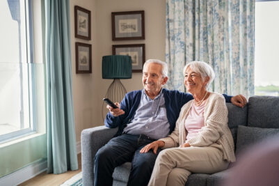 Happy senior couple in living room using remote control to change channel. Seniors watch TV for entertainment while man using remote control. Cheerful wife and old man sitting on couch in living room with television show.